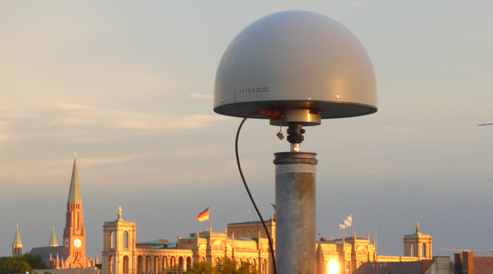 SAPOS-Referenzstationsantenne auf dem Dach des Münchner LDBVs. Im Hintergrund der bayerische Landtag und die St. Johann Baptist Kirche am Johannisplatz.